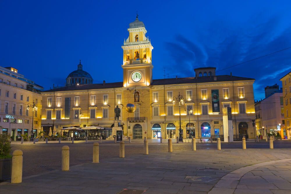 Piazza Garibaldi a Parma di sera, con il Palazzo del Governatore illuminato e la torre dell'orologio sotto un cielo blu intenso.
