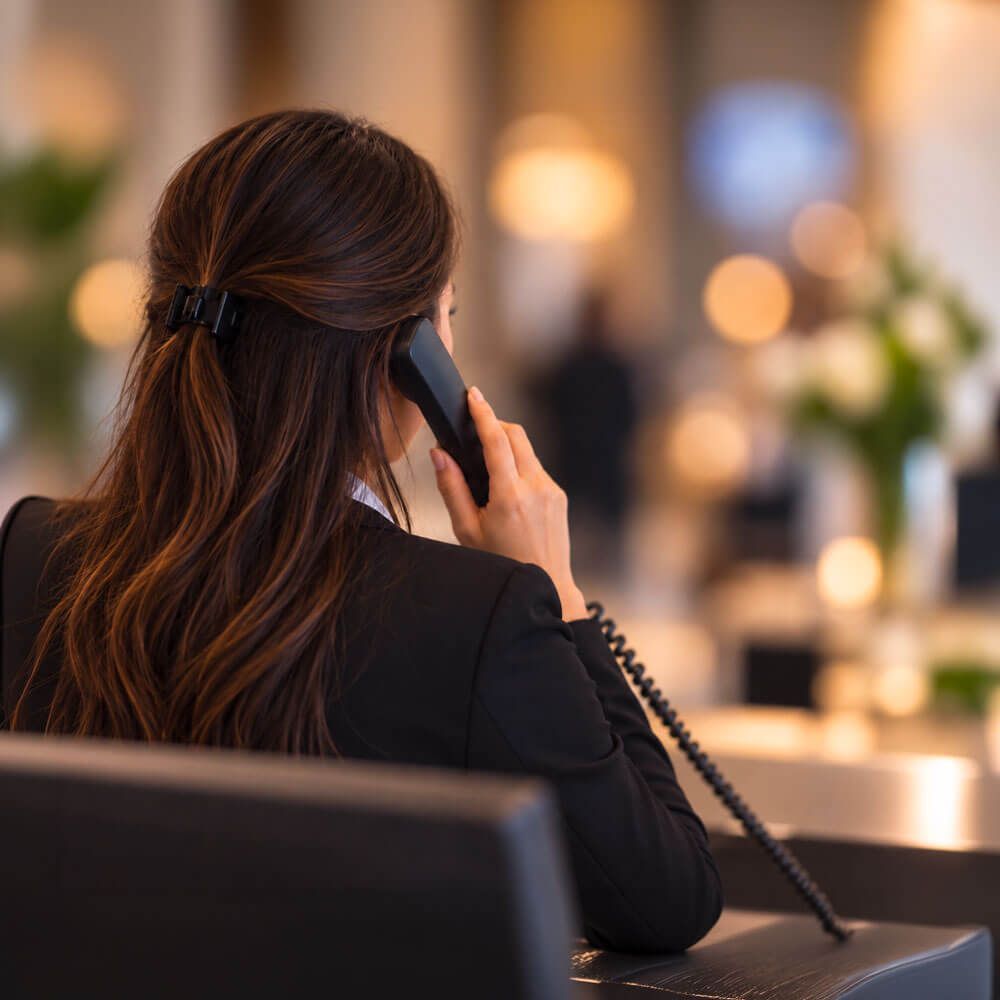 Receptionist in giacca scura, di spalle, al telefono con un apparecchio fisso nella hall dell'hotel.
