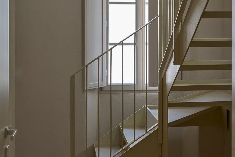 Scala interna con pareti bianche e ringhiera in metallo, illuminata dalla luce naturale di un'ampia finestra.