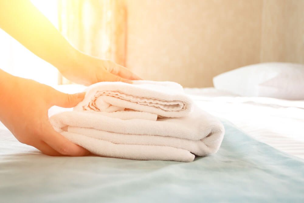 Mani che sistemano una pila di asciugamani bianchi piegati su un letto, in una stanza luminosa.