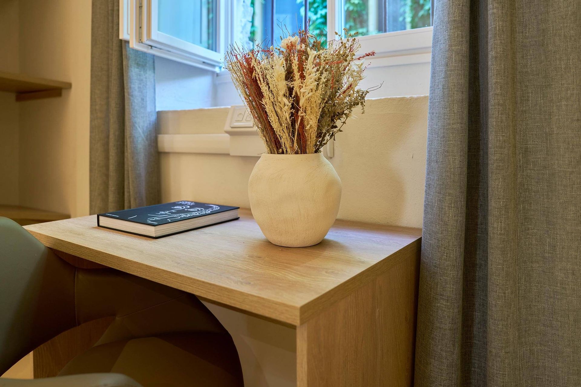 Vaso in ceramica chiara con fiori secchi nei toni del beige e del ruggine, su una scrivania in legno accanto a una finestra.