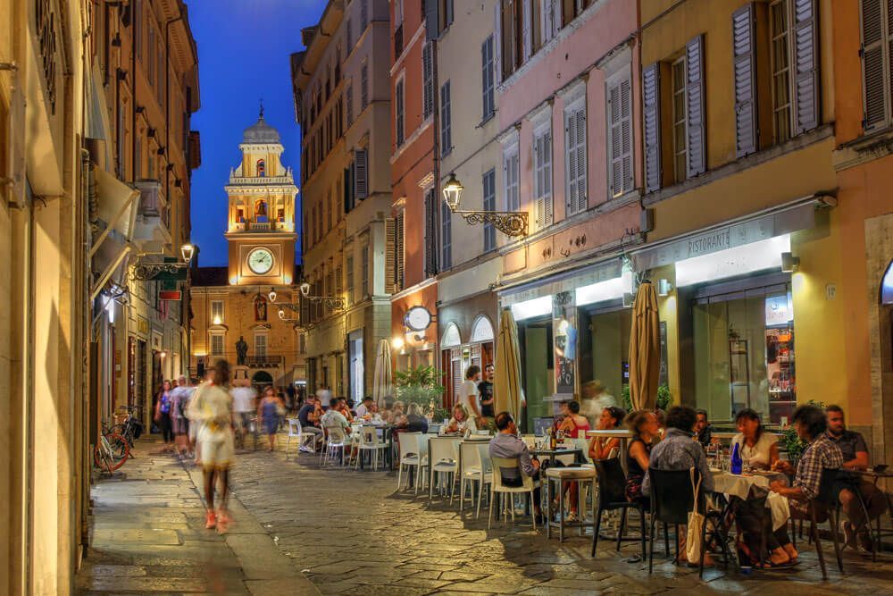 Via del centro storico di Parma al tramonto, con tavolini all'aperto affollati davanti ai caffè e, sullo sfondo, la torre dell'orologio del Palazzo del Governatore illuminata.