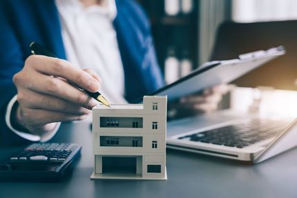Ein Mann sitzt an einem Schreibtisch und hält einen Stift und ein Modellhaus in der Hand.