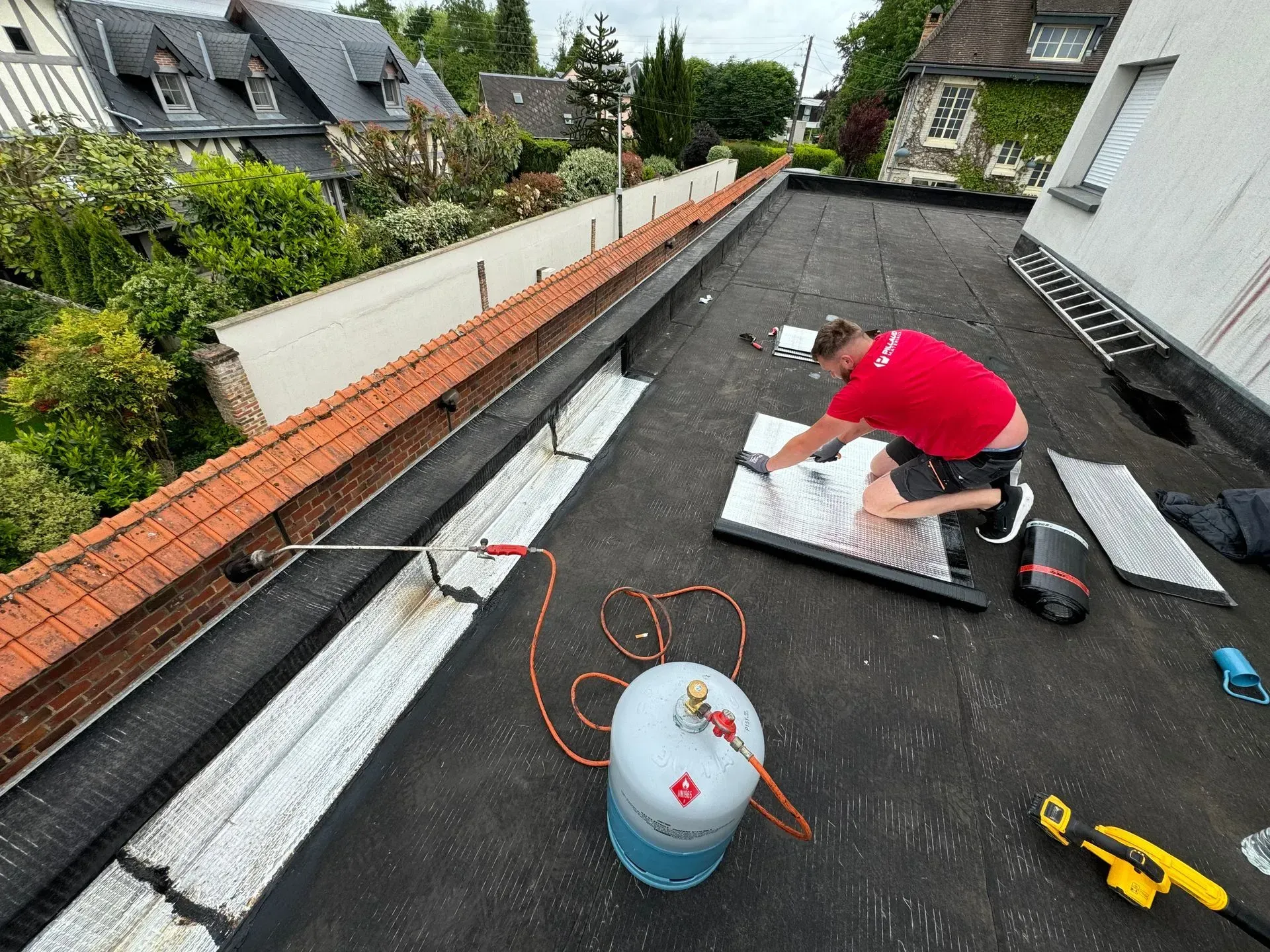 Un ouvrier en chemise installe une membrane d'étanchéité sur un toit plat, avec ses outils et matériaux à proximité.