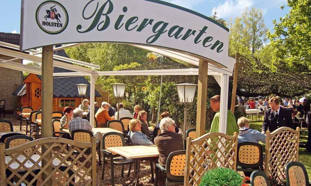 Ein sonniger und gut besuchter Biergarten im Freien, in dem unter einem weißen Torbogen Tische mit Gästen besetzt sind.