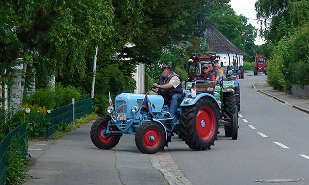 Ein blauer Traktor zieht einen roten Traktor auf einer von Bäumen gesäumten Straße; in den Fahrzeugen befinden sich Personen.