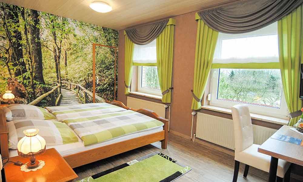 Helles Schlafzimmer mit Bett, Waldmotiv an der Wand, grünen Akzenten und einer Sitzecke am Fenster
