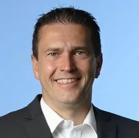 Lächelnder Mann in dunklem Anzug und weißem Hemd vor hellblauem Hintergrund