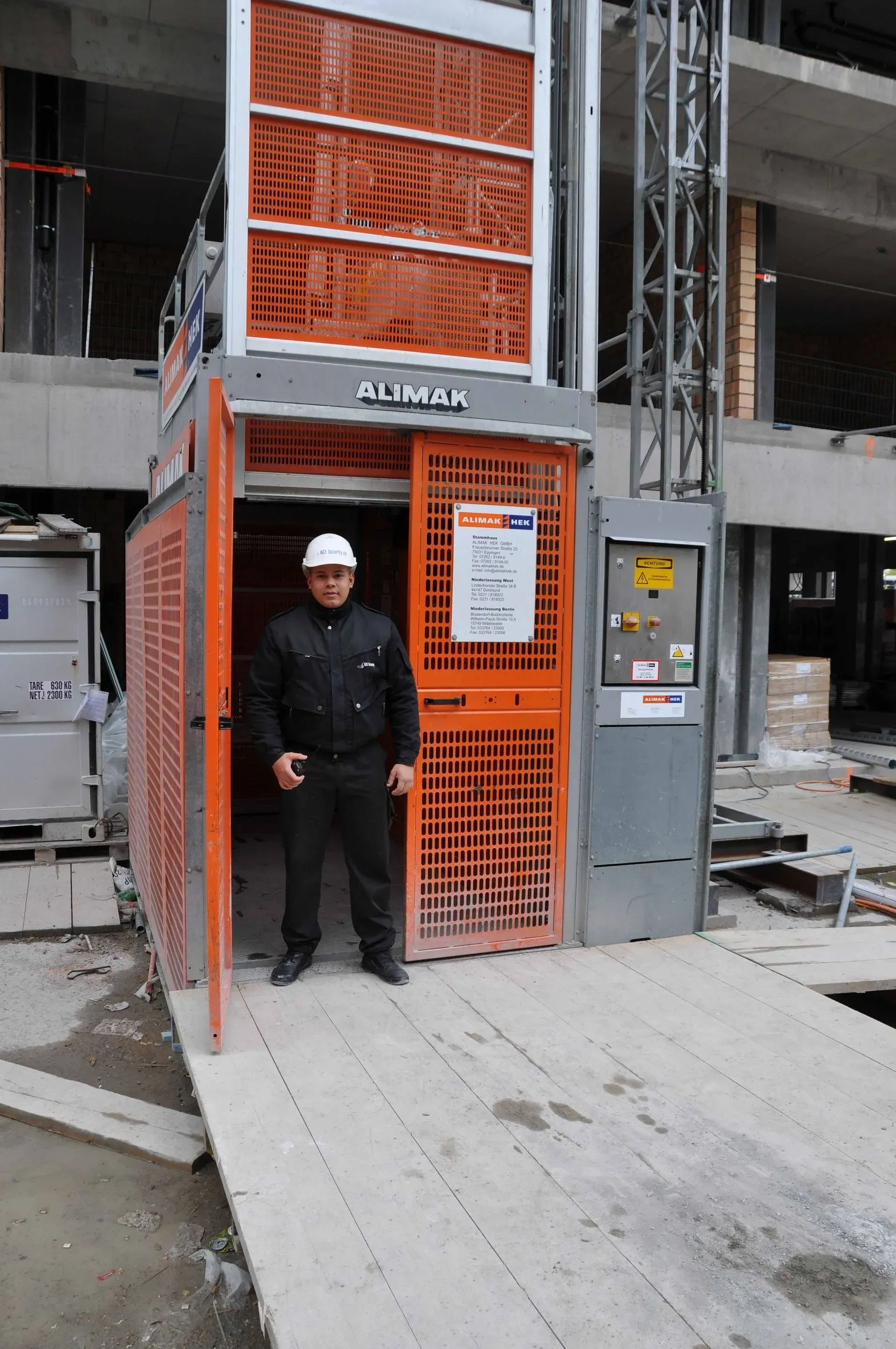 Ein Arbeiter mit weißem Schutzhelm steht neben einem orangefarbenen Bauaufzug auf einer Baustelle.