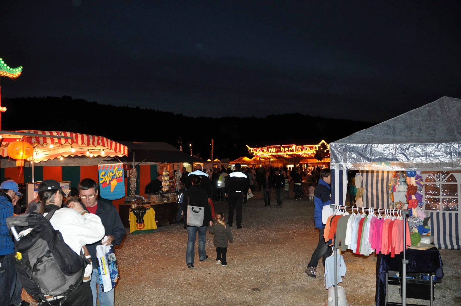 Nächtlicher Freiluftmarkt mit beleuchteten Ständen und Menschen, die zwischen Zelten und Buden umhergehen.