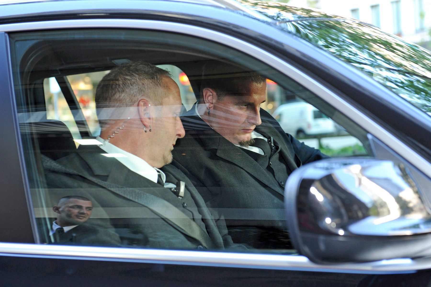 Zwei Männer in dunklen Mänteln sitzen in einem Auto, das durch das Seitenfenster zu sehen ist.