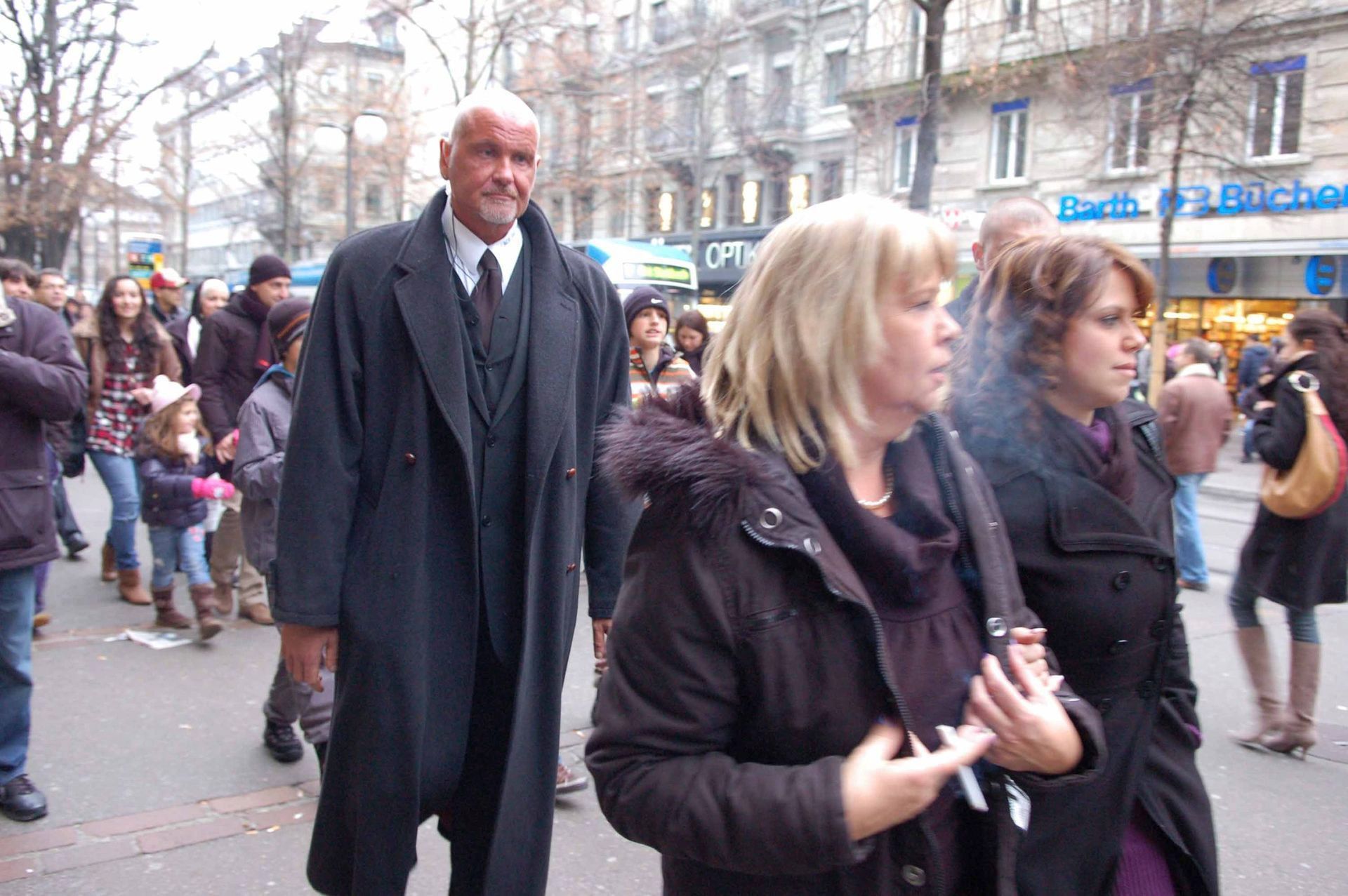 Menschenmenge, die eine Stadtstraße entlanggeht; im Vordergrund drei Personen, im Hintergrund Gebäude.