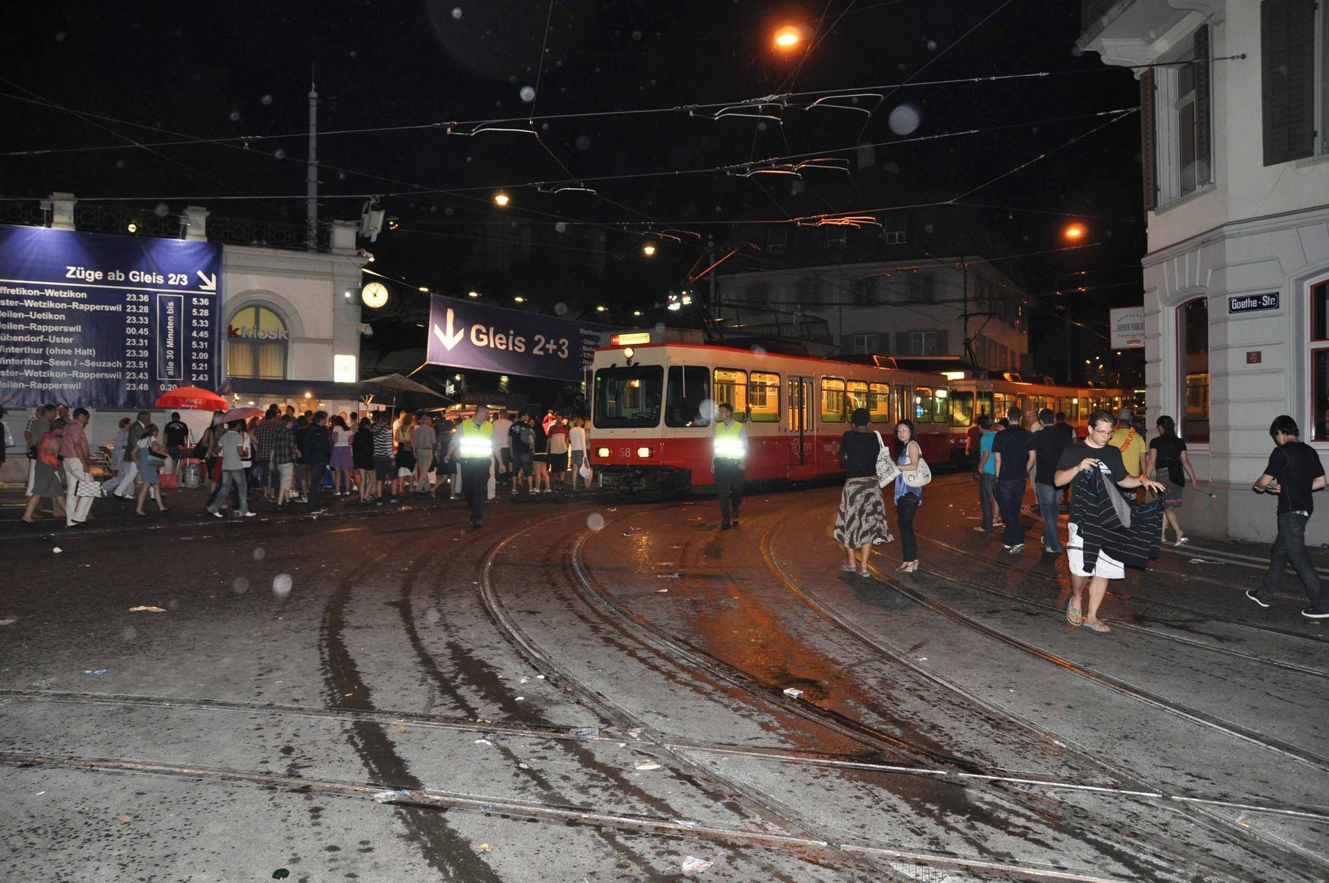 Nächtliche Straßenszene mit einer roten Straßenbahn auf nassen Gleisen, Menschenmengen auf den Bürgersteigen