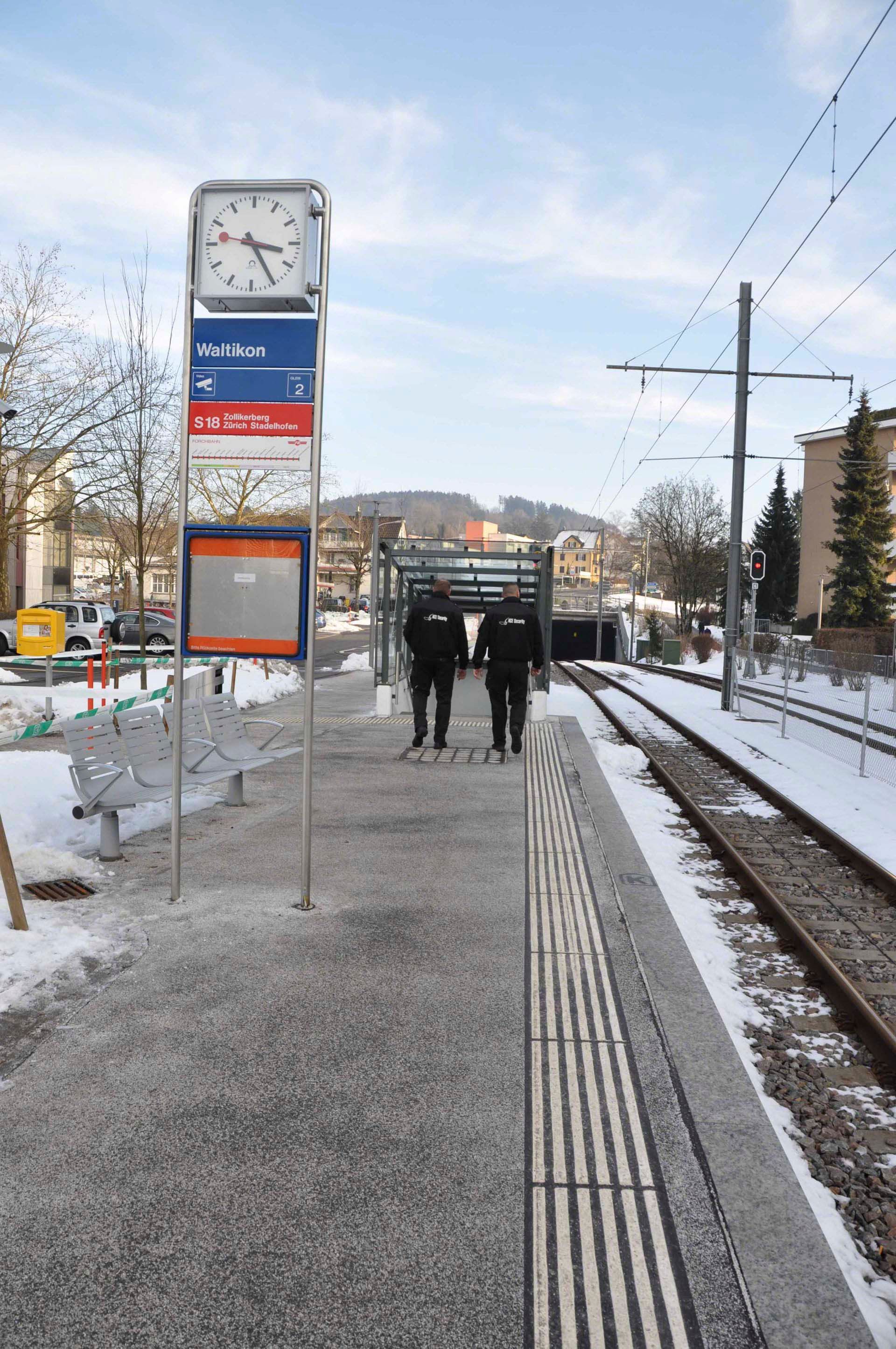 Verschneite Straßenbahnhaltestelle mit zwei Personen, die auf dem Bahnsteig neben den Gleisen entlanggehen