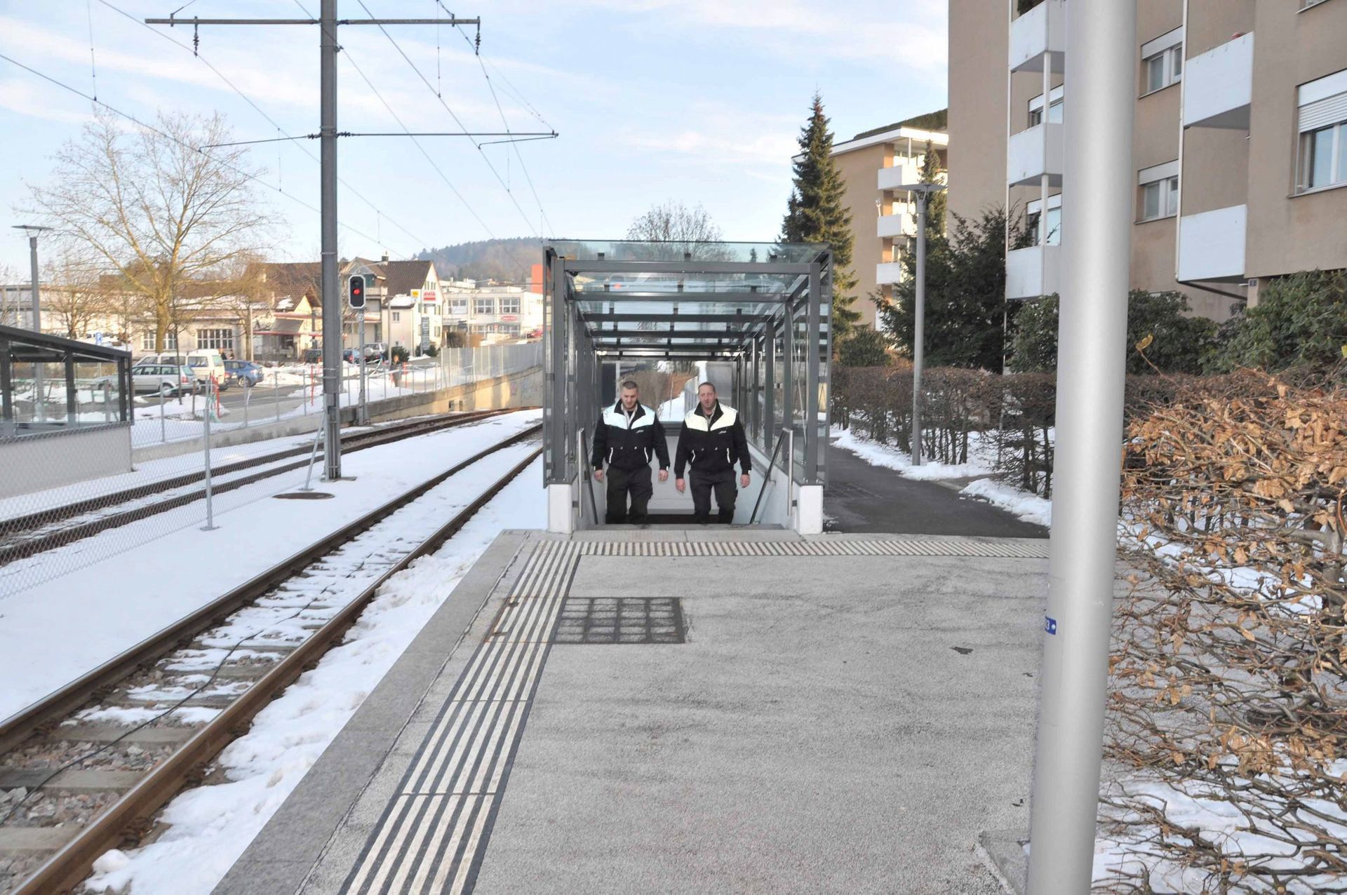 Menschen warten auf einem verschneiten Bahnsteig mit einem überdachten Wartehäuschen und nahegelegenen Gleisen.
