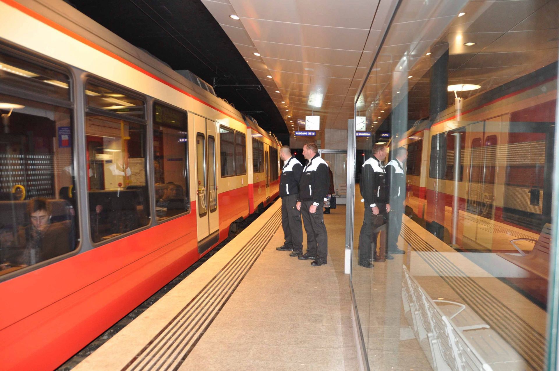 Bahnsteig mit einem rot-weißen Zug und drei uniformierten Mitarbeitern, die in der Nähe von Glastüren stehen