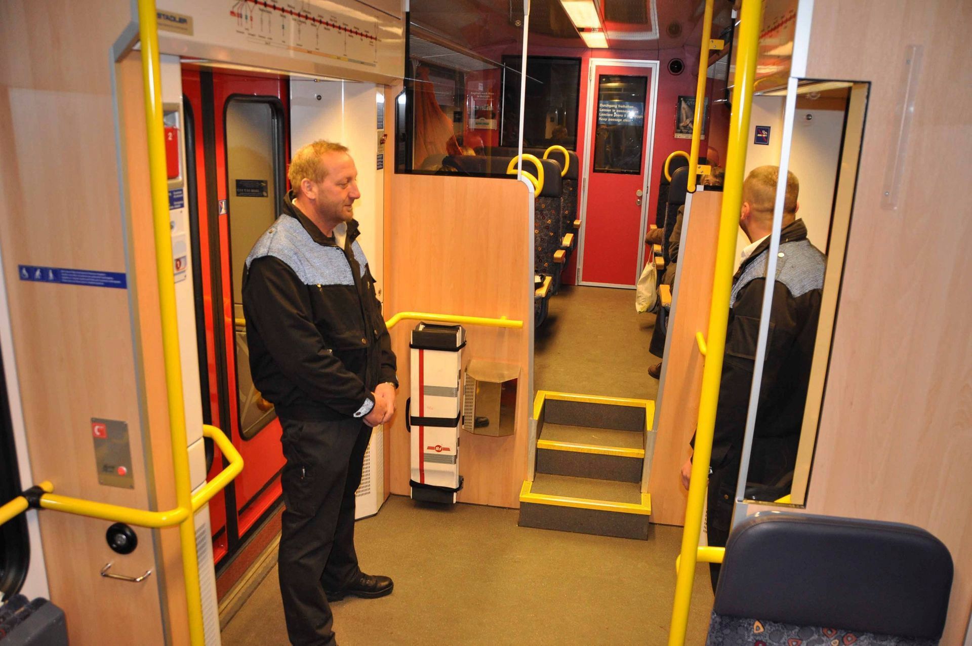 Ein Mann steht in einem Zugwaggon mit gelben Handläufen und roten Türen und blickt zum Gang.