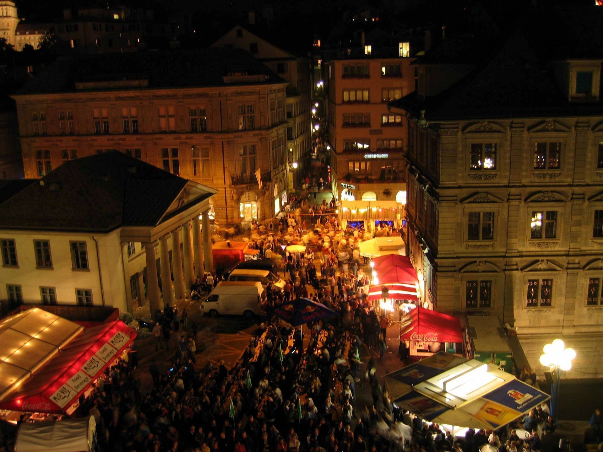 Ein geschäftiges nächtliches Straßenfest zwischen beleuchteten Gebäuden, mit Zelten und warmen orangefarbenen Lichtern.