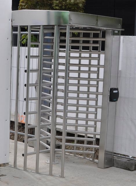 Drehkreuz aus Metall an einem Außeneingang mit weißen Zaunelementen und einem Tastenfeld auf der rechten Seite