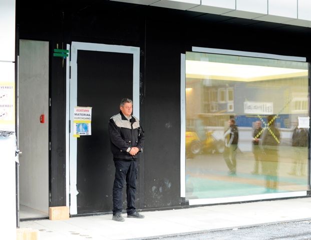 Ein Mann steht vor einem dunklen Ladeneingang, neben einem großen Fenster, in dem sich die Menschen im Inneren spiegeln.
