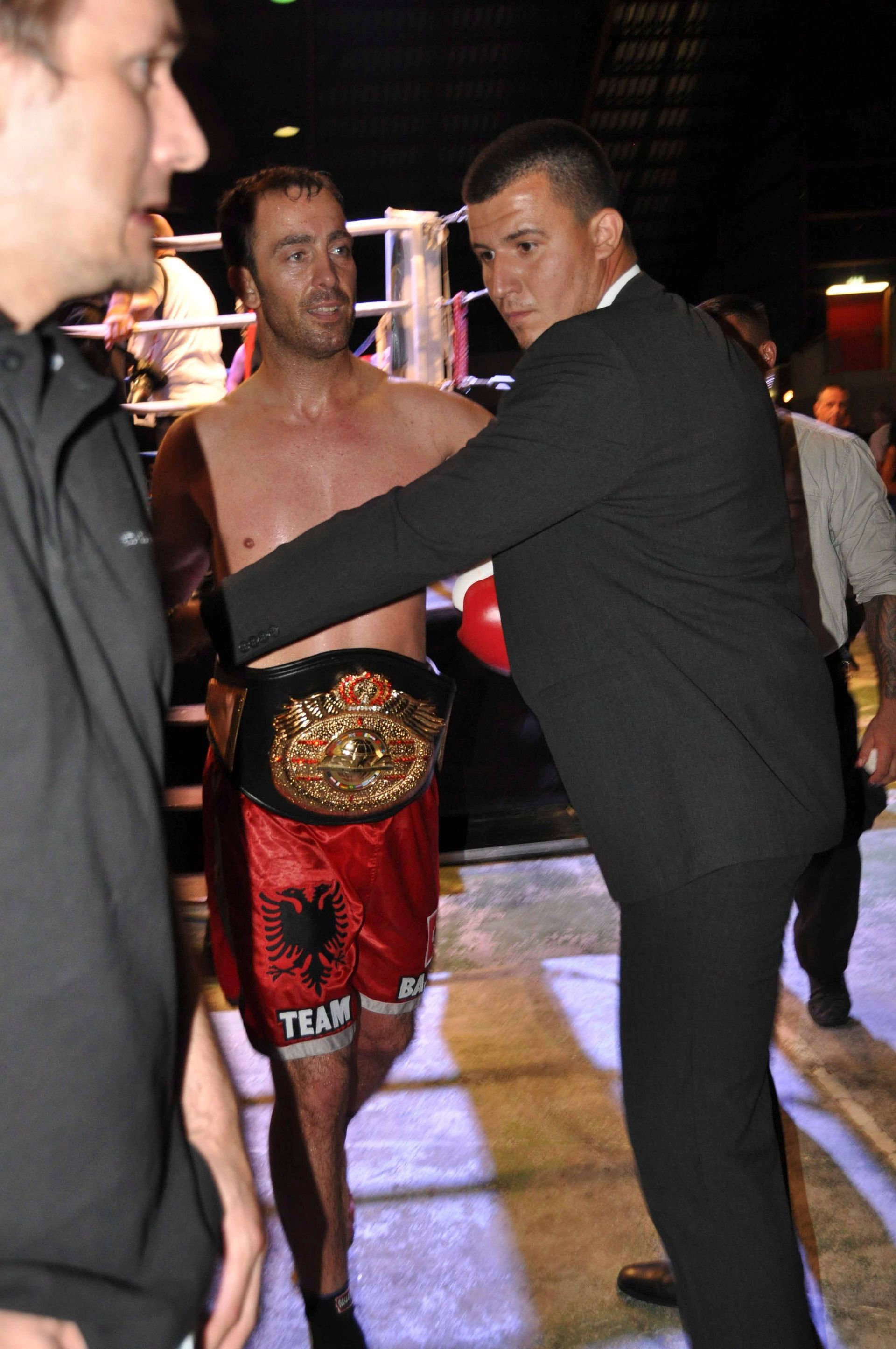 Ein Mann im schwarzen Anzug richtet einen oberkörperfreien Boxer, der einen Meisterschaftsgürtel trägt, in einem Boxring.