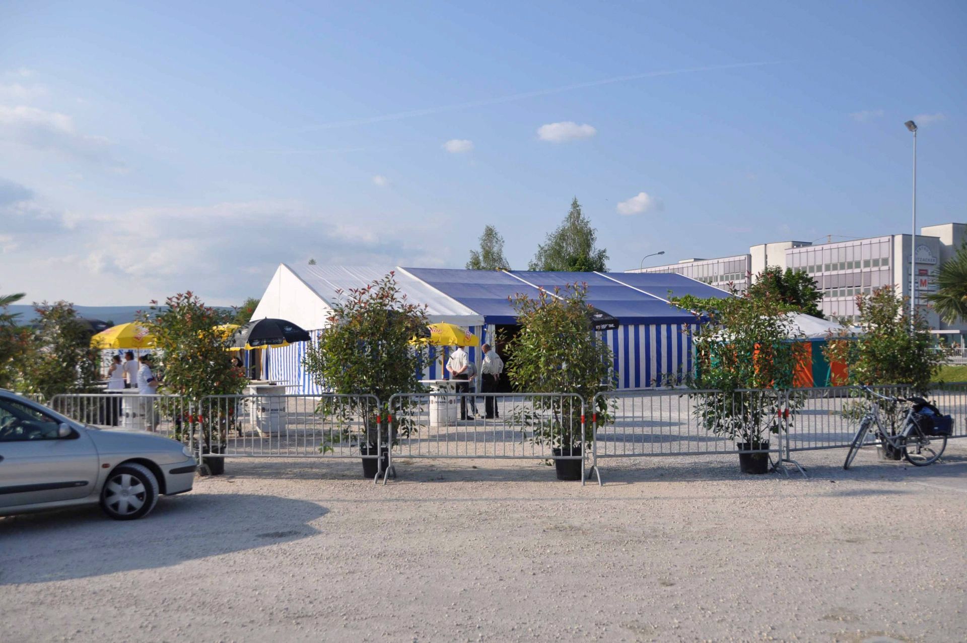 Modernes Gebäude mit Solarpaneelen hinter einem Zaun, dazwischen parkende Autos und Fahrräder unter blauem Himmel.