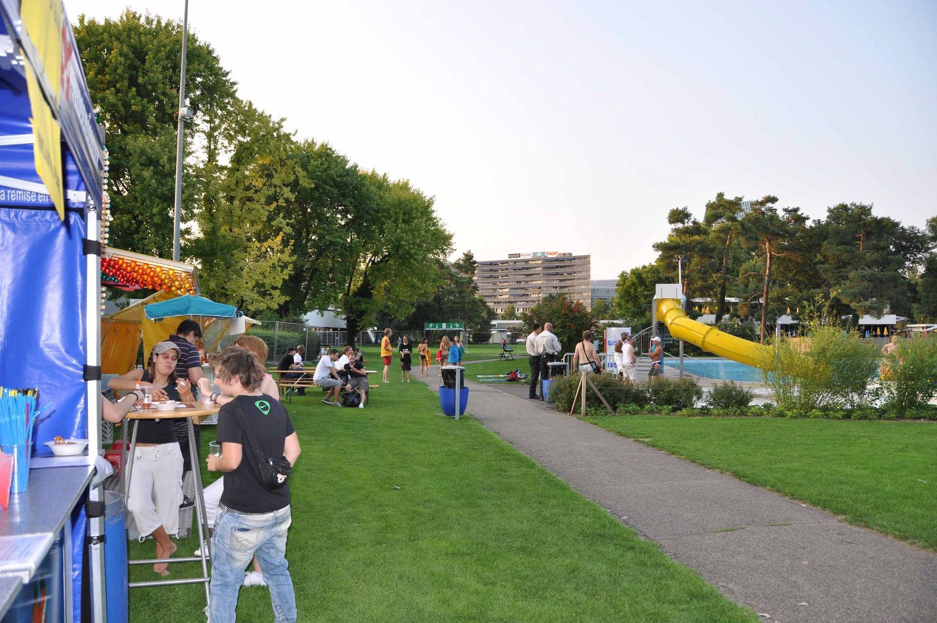 Menschen auf einem Jahrmarkt im Park; im Hintergrund sind Stände, Bäume, ein Weg und eine gelbe Rutsche zu sehen.