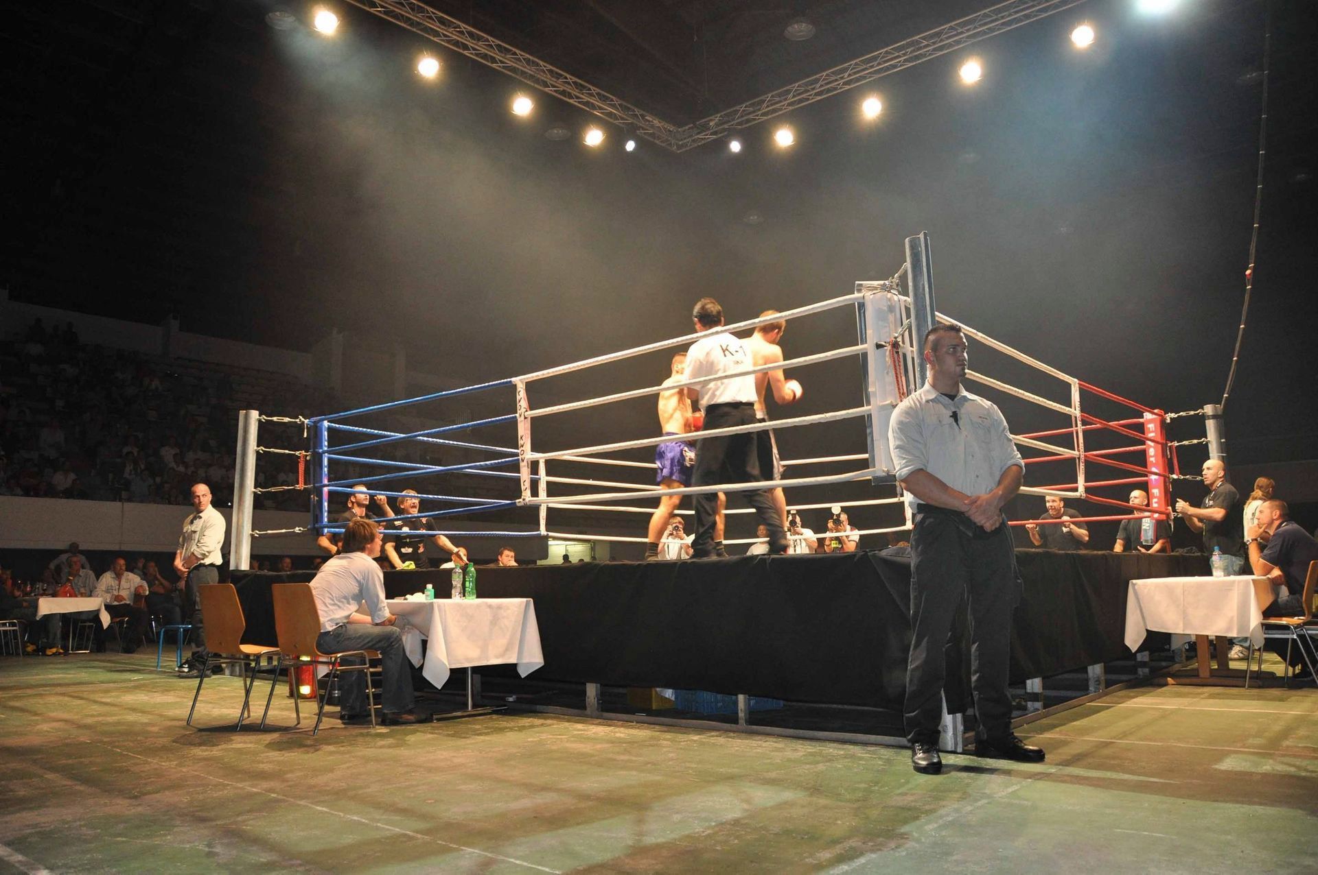 Boxring unter hellem Licht, Kämpfer und Ringrichter in Aktion, Publikum rund um die Arena.