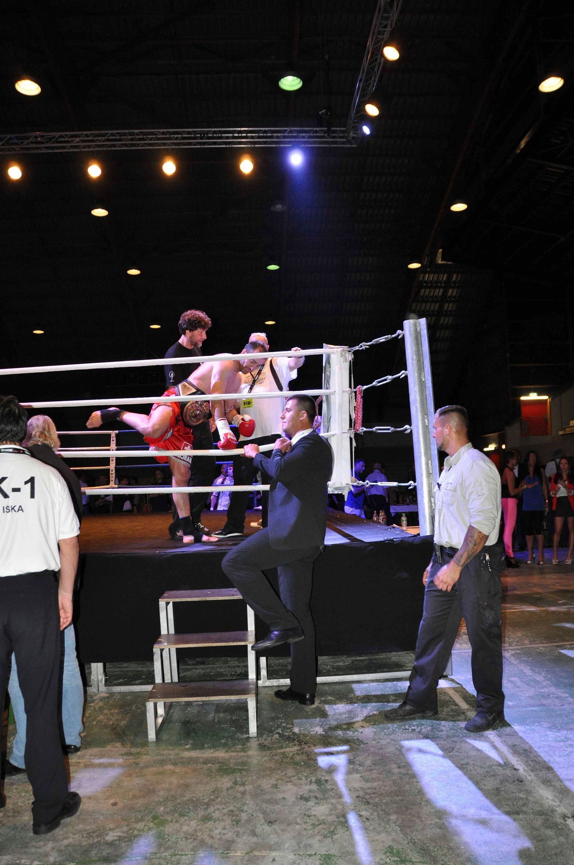 Boxring mit zwei Kämpfern und Offiziellen in einer schwach beleuchteten Arena, einer klettert die Seile hoch.