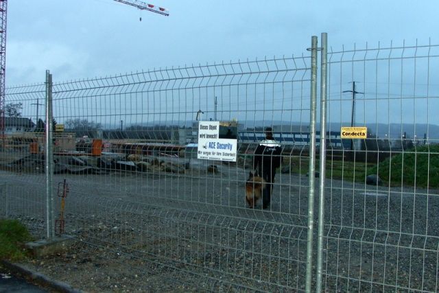 Maschendrahtzaun auf einer Baustelle mit Geräten und Warnschildern unter bewölktem Himmel