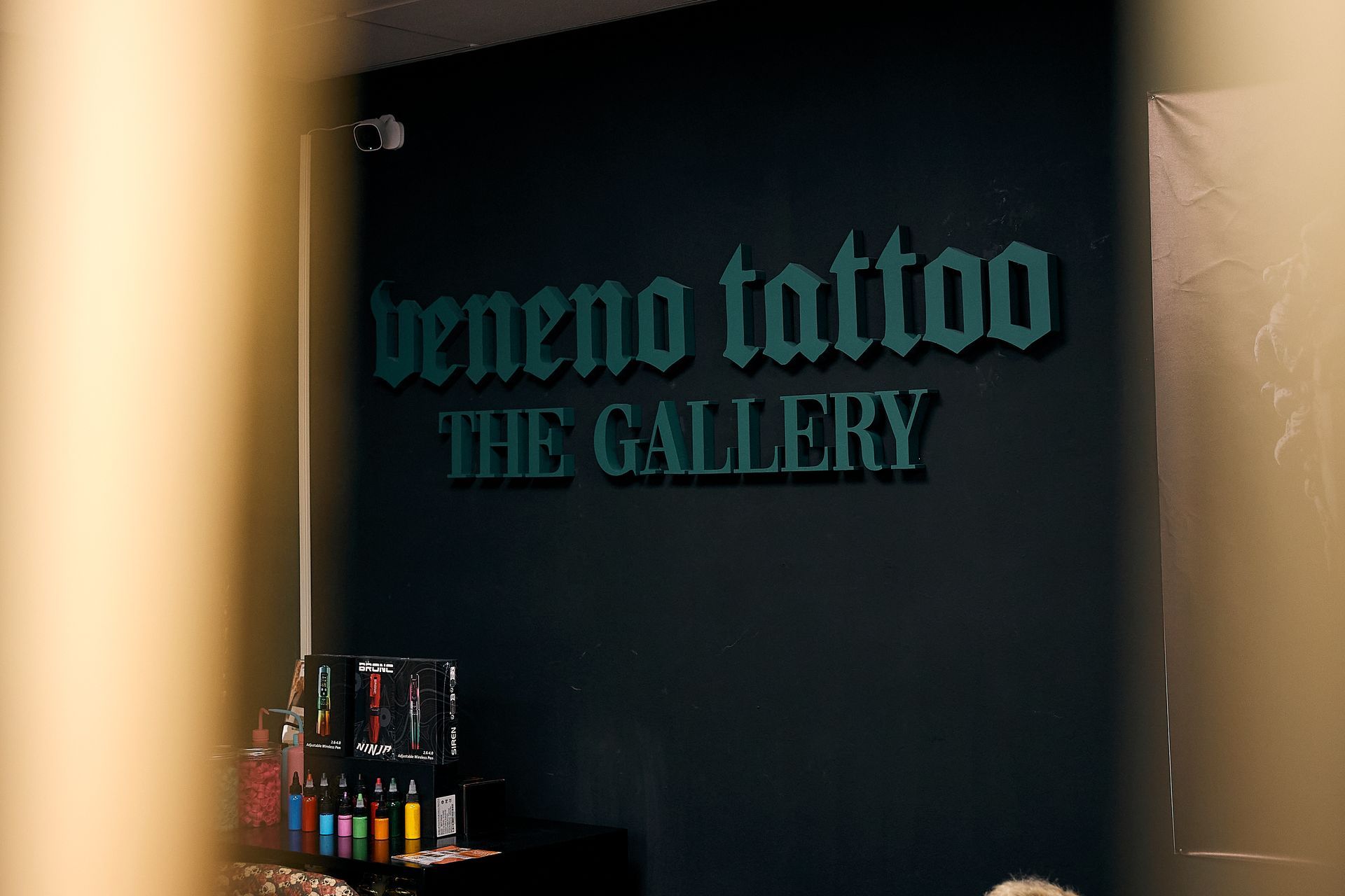 Pared oscura con las letras “bene no tto” y “THE GALLERY” en color verde azulado, iluminada por una luz cálida a la izquierda.