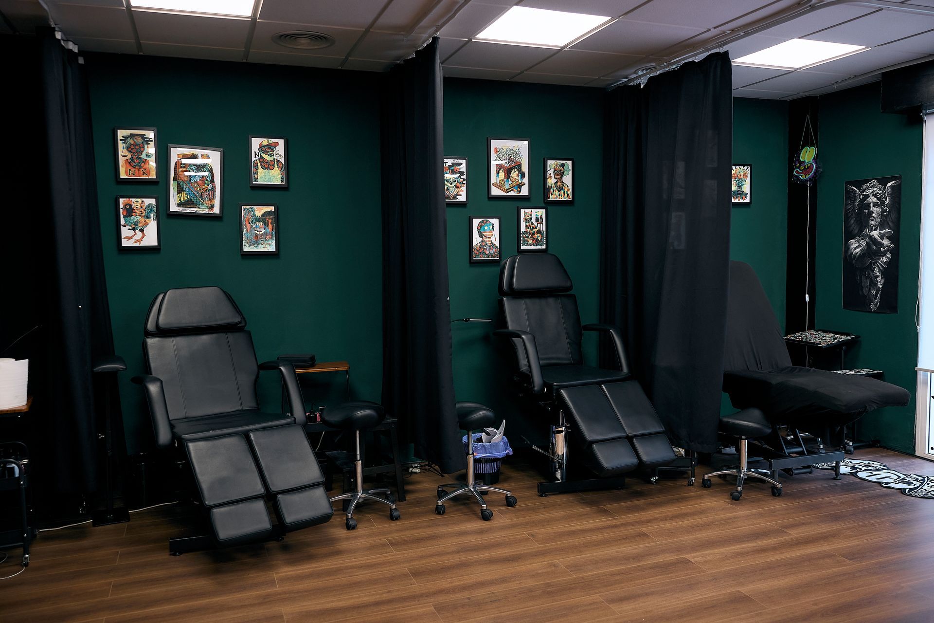 Estudio de tatuajes con dos sillas negras, paredes verdes, cuadros enmarcados y suelo de madera.