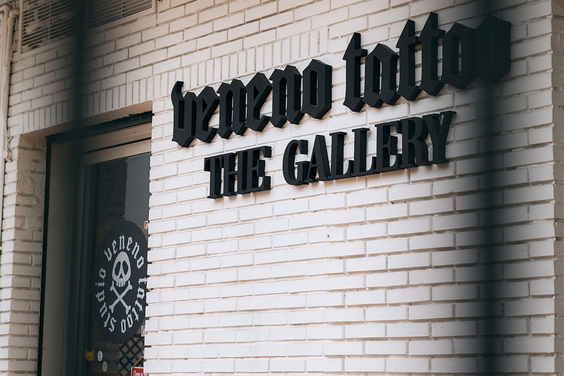 Pared exterior de ladrillo con un letrero negro que dice “bechto tattoo THE GALLERY” encima de una puerta.