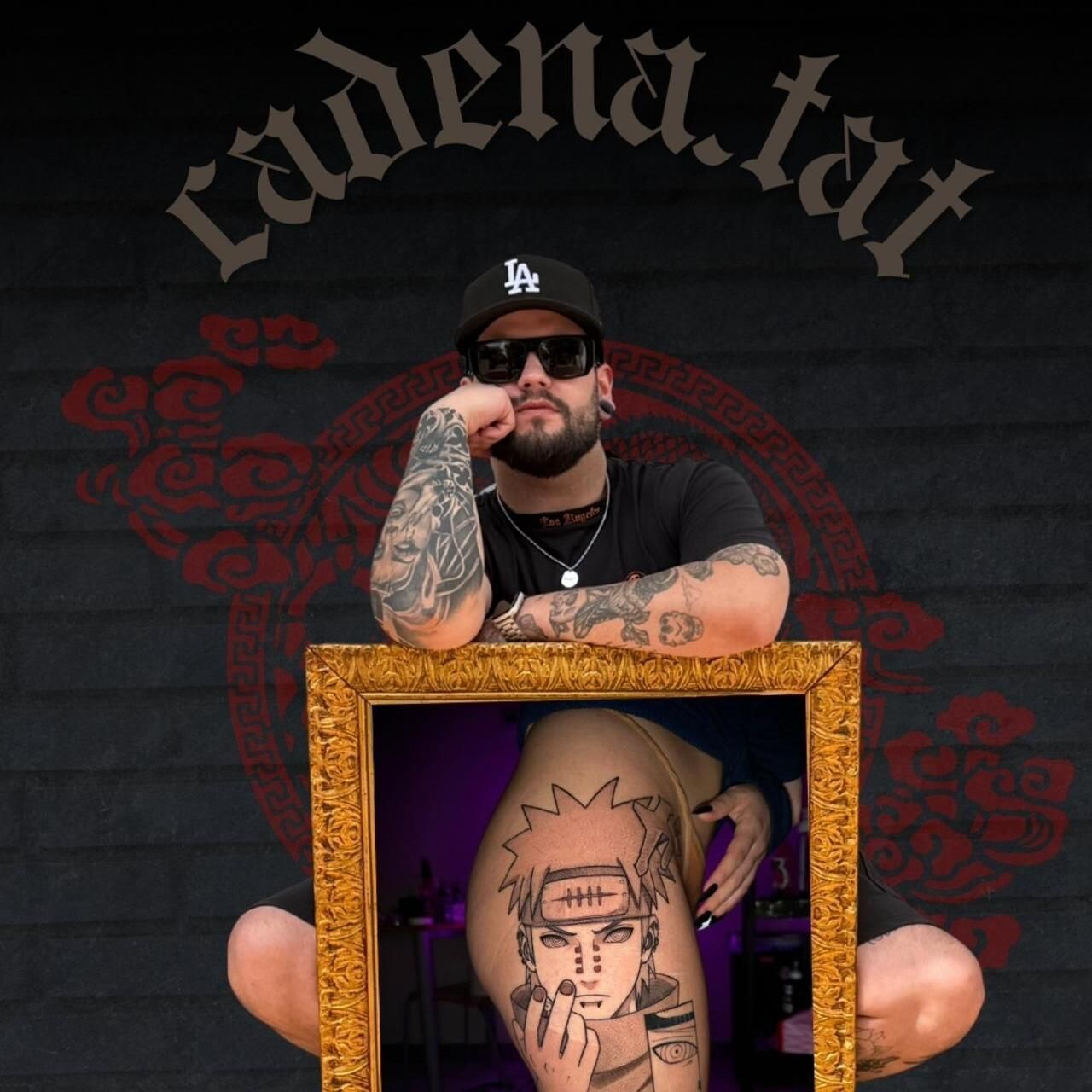 Un hombre con gafas de sol y gorra está sentado frente a un diseño oscuro estilo tatuaje, sosteniendo un retrato enmarcado de un rostro.
