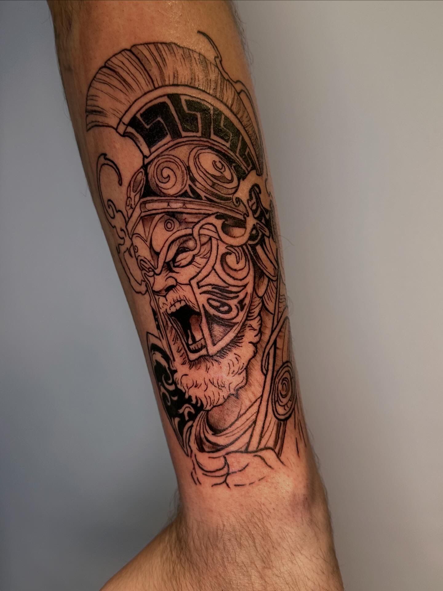 Un detallado tatuaje en blanco y negro de un guerrero espartano gruñendo, con un casco corintio de estilo griego, en la parte inferior de la pierna.