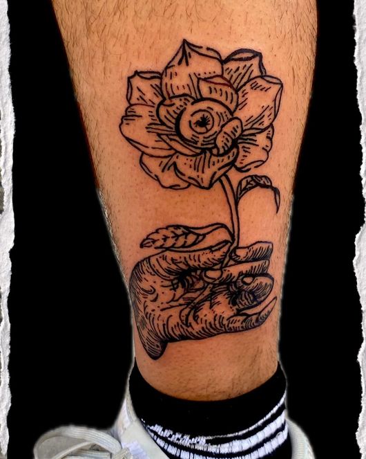 Tatuaje floral en blanco y negro en una pantorrilla, con una flor en plena floración que brota de una pequeña mano que sostiene una cara.
