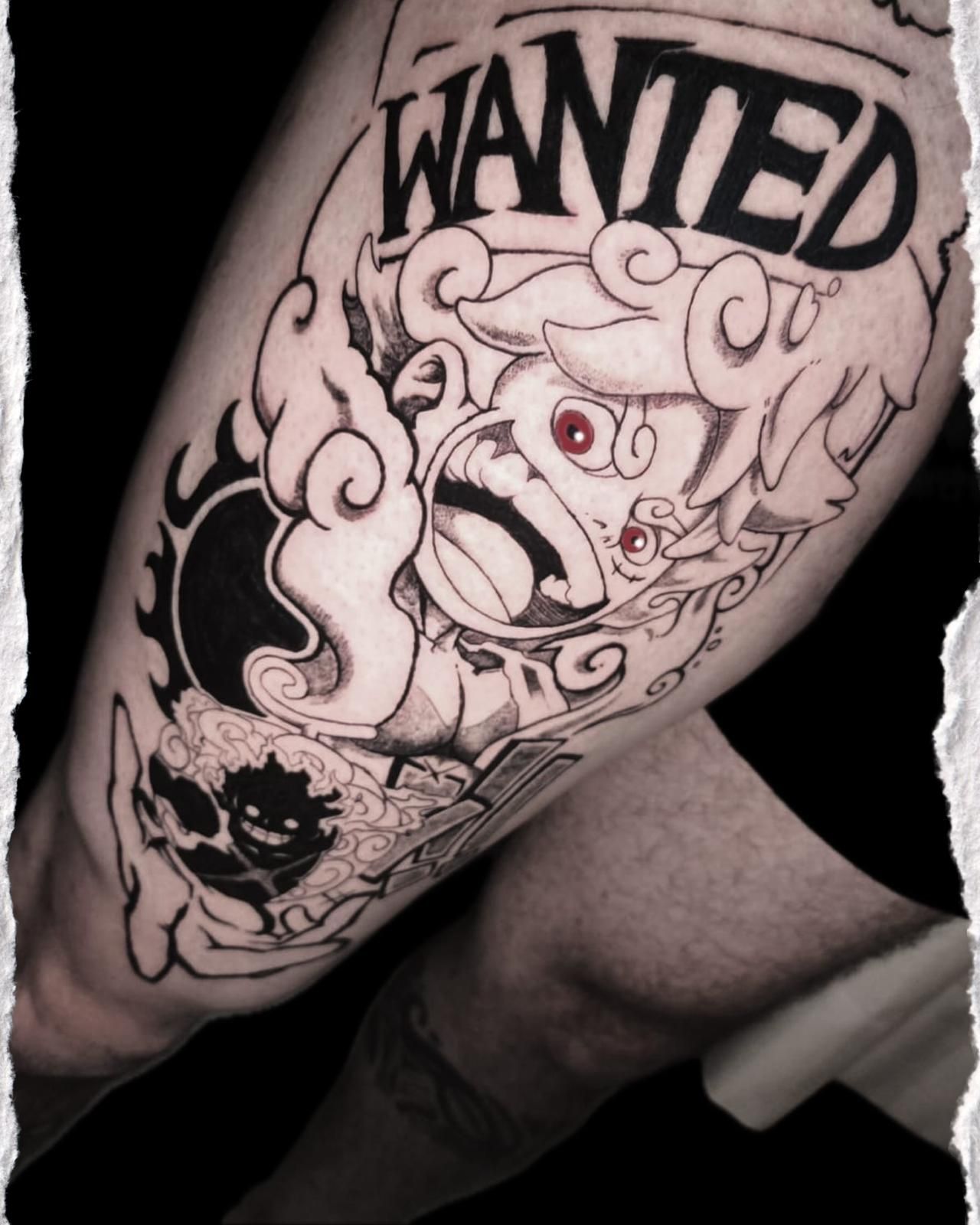 Tatuaje en blanco y negro en un brazo doblado que muestra una figura de dibujos animados con la palabra "SE BUSCA" encima.
