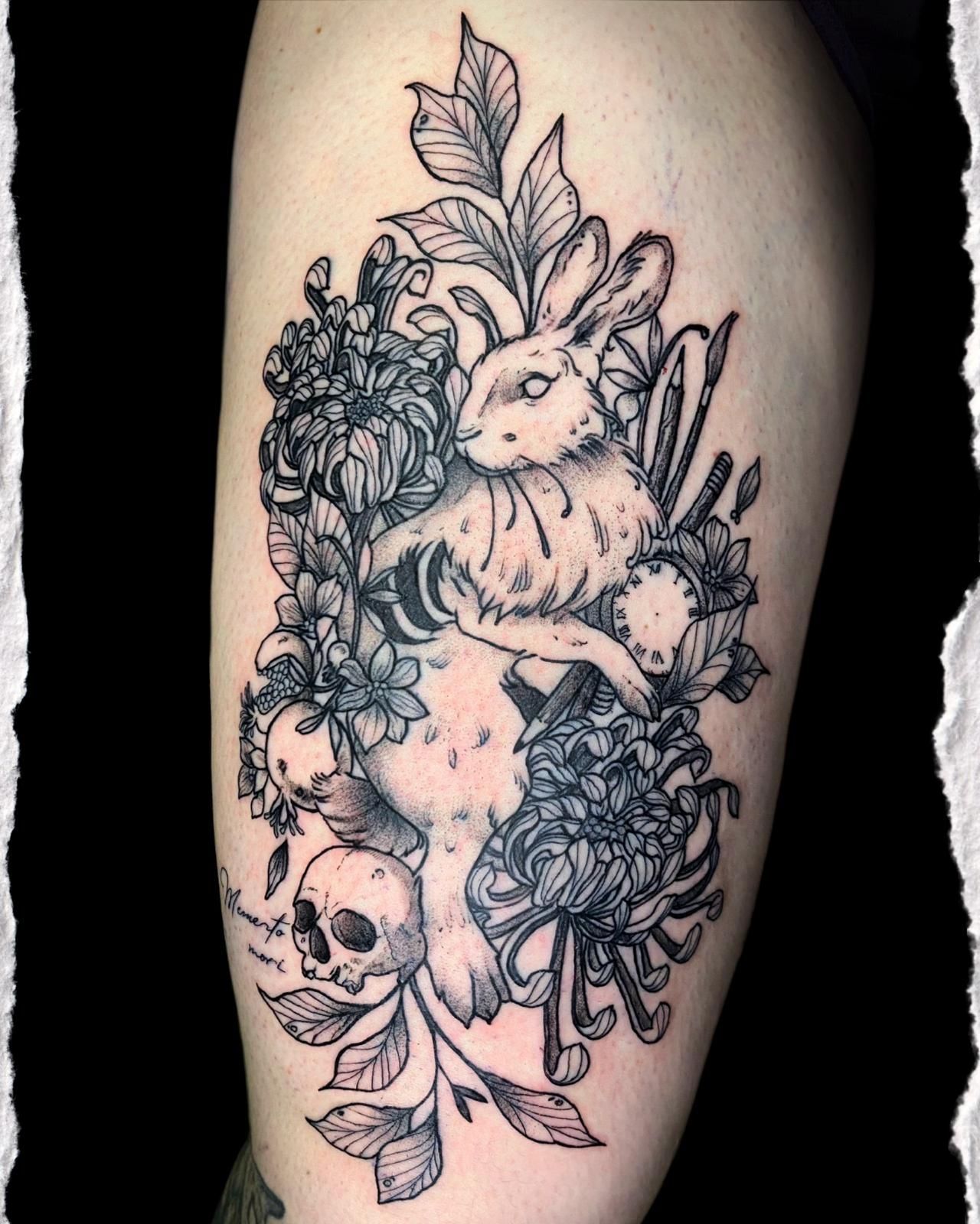Tatuaje en blanco y negro de un conejo rodeado de flores y hojas en el antebrazo.