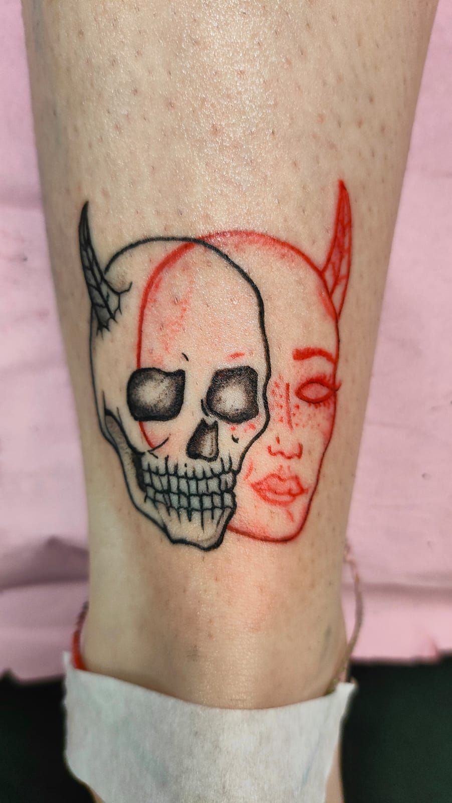 Un tatuaje en una pierna que muestra una calavera en tinta negra superpuesta a un rostro en tinta roja con cuernos y pecas.