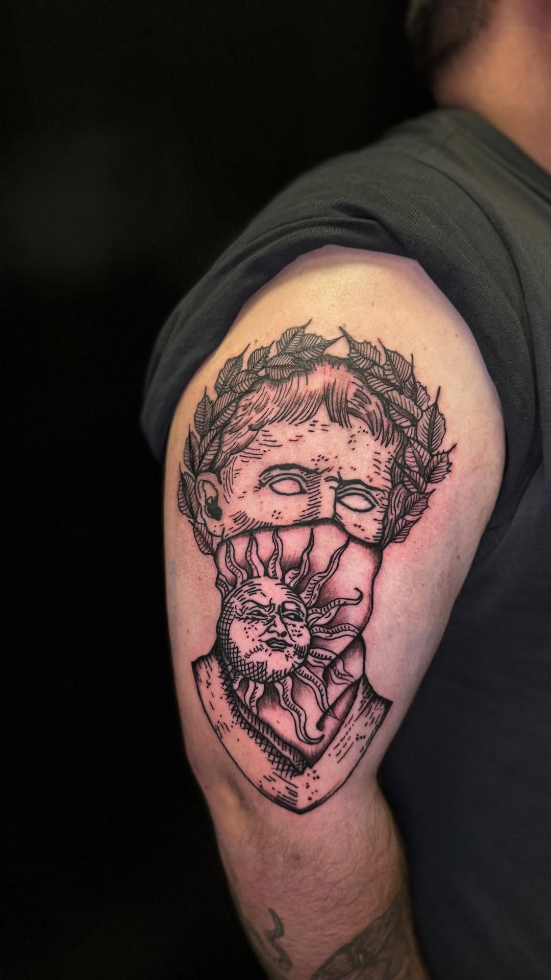 Un tatuaje de un busto romano con una corona de laurel y una máscara decorada con un rostro solar.
