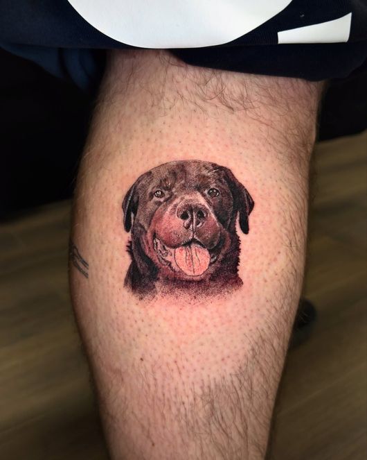 Pequeño tatuaje de un perro negro y marrón en la pantorrilla de una persona, con la lengua del perro fuera.