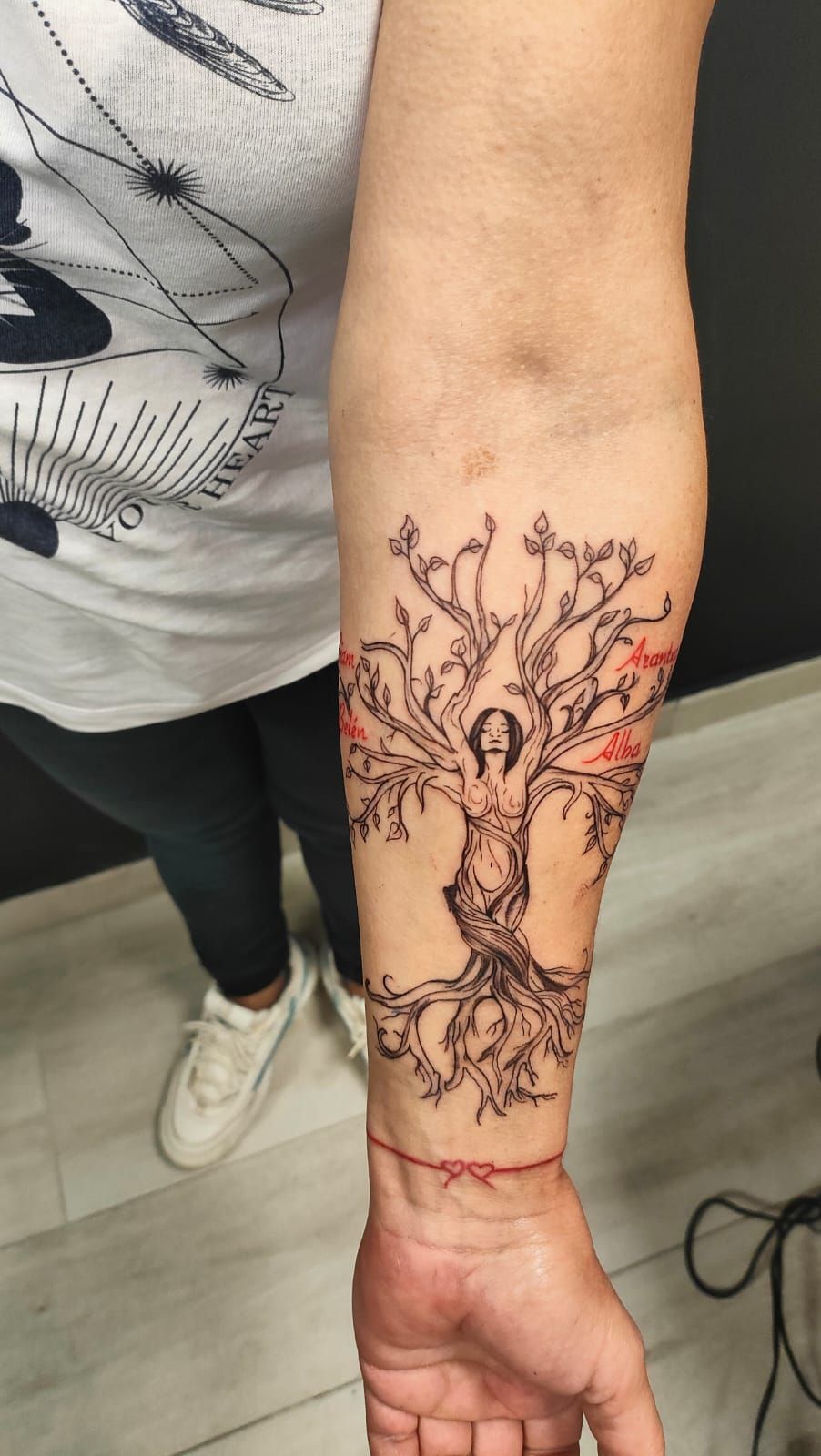 Brazo con un tatuaje de un árbol dibujado con líneas negras y una muñequera roja, extendido sobre un suelo gris.