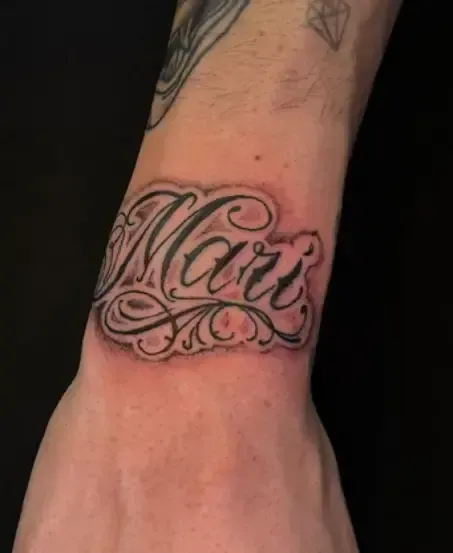 Tatuaje en la muñeca con una caligrafía ornamentada en tinta negra sobre piel clara.