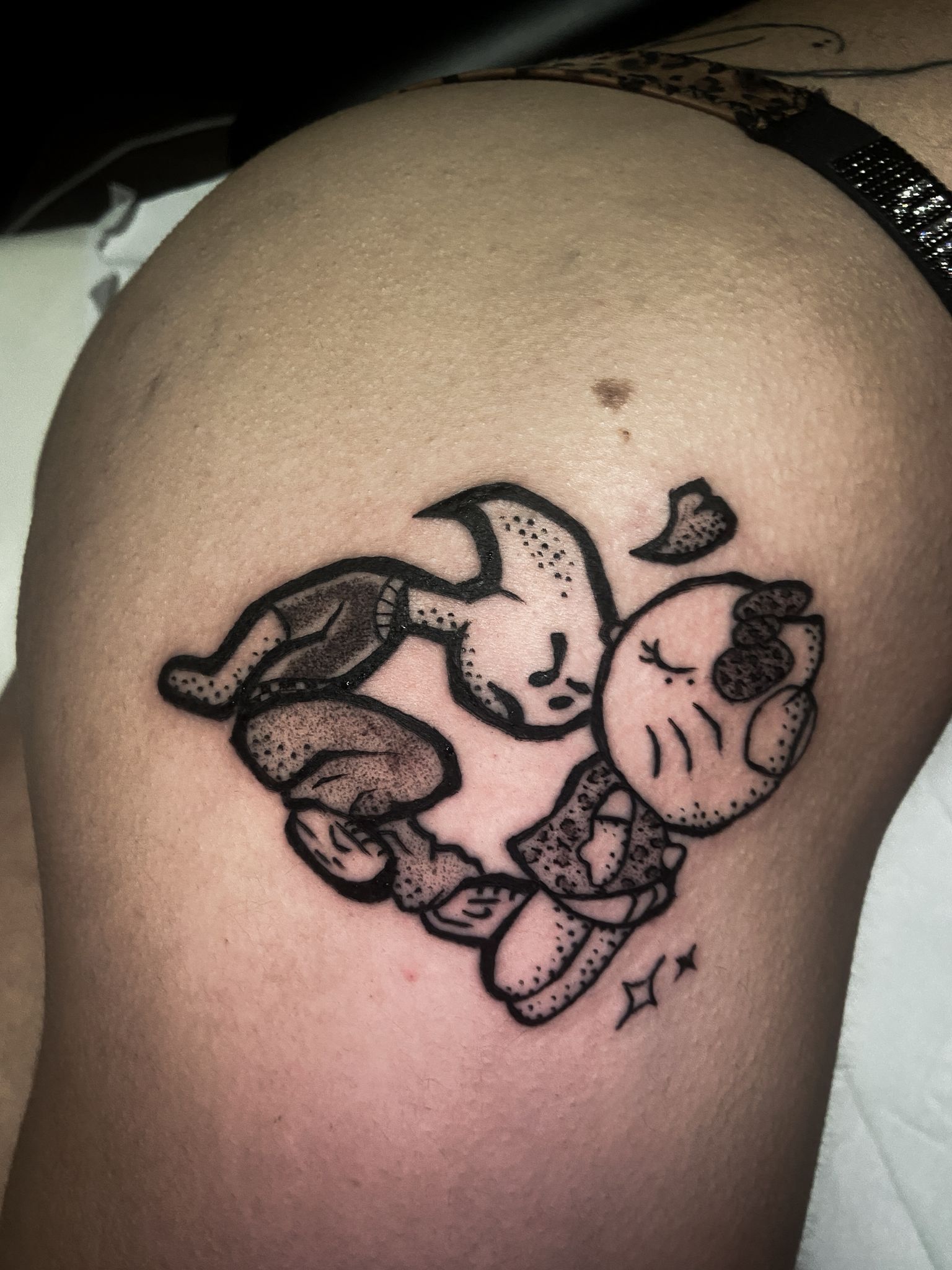 Un tatuaje en tinta negra sobre la piel que representa dos figuras de dibujos animados formando un corazón, con sombreado punteado y pequeños detalles en forma de estrella.