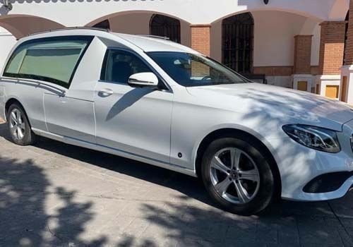 Un coche fúnebre Mercedes Benz de color blanco está estacionado frente a un edificio.