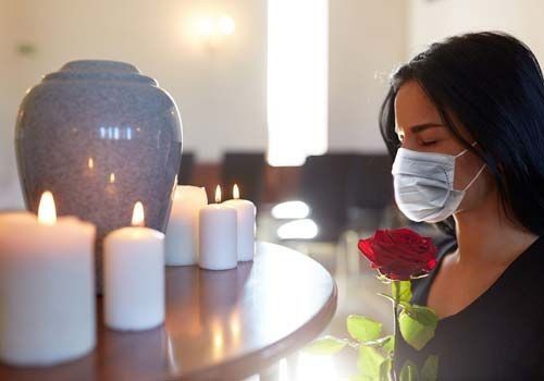 Una mujer que lleva una mascarilla sostiene una rosa delante de una urna y velas.