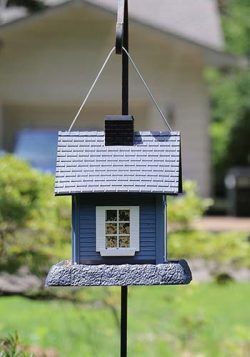 Petit nichoir bleu et blanc suspendu à l'extérieur par une tige noire, avec un arrière-plan de jardin flou.