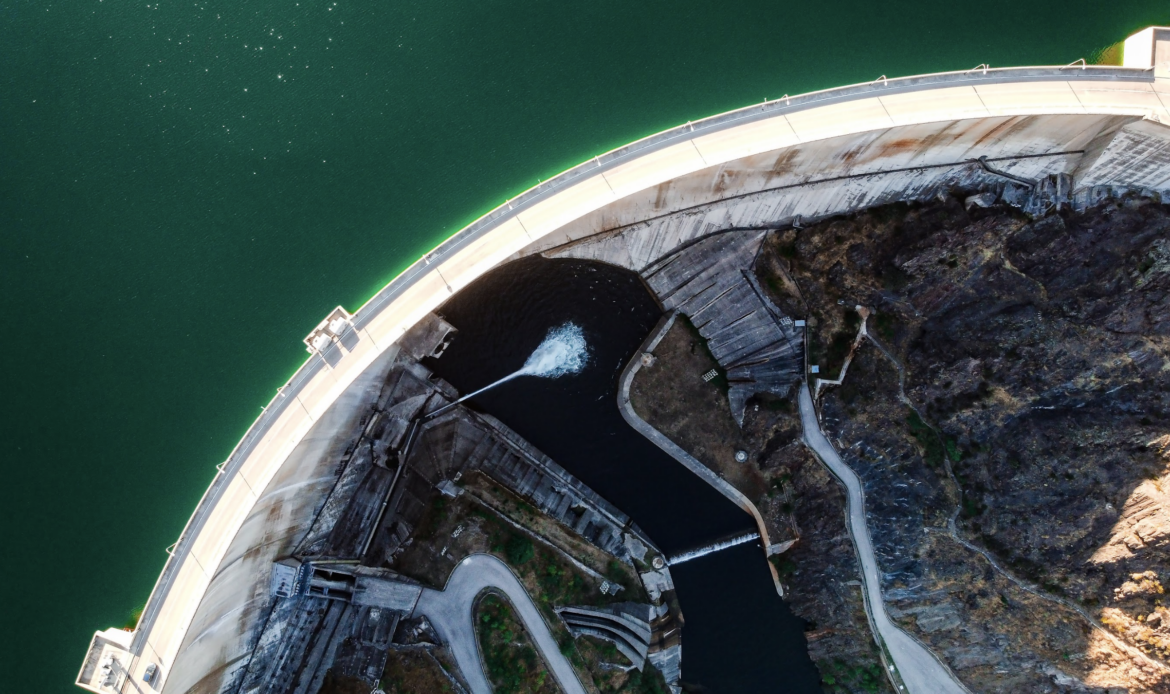 Vista aérea de una gran presa de hormigón sobre un lago verde. El agua salpica desde la presa, con una carretera a un lado.