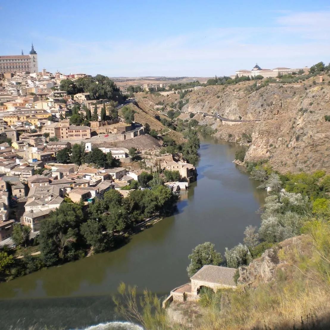 Ciudad de Toledo, España, con edificios en una colina junto a un río bajo un cielo soleado.