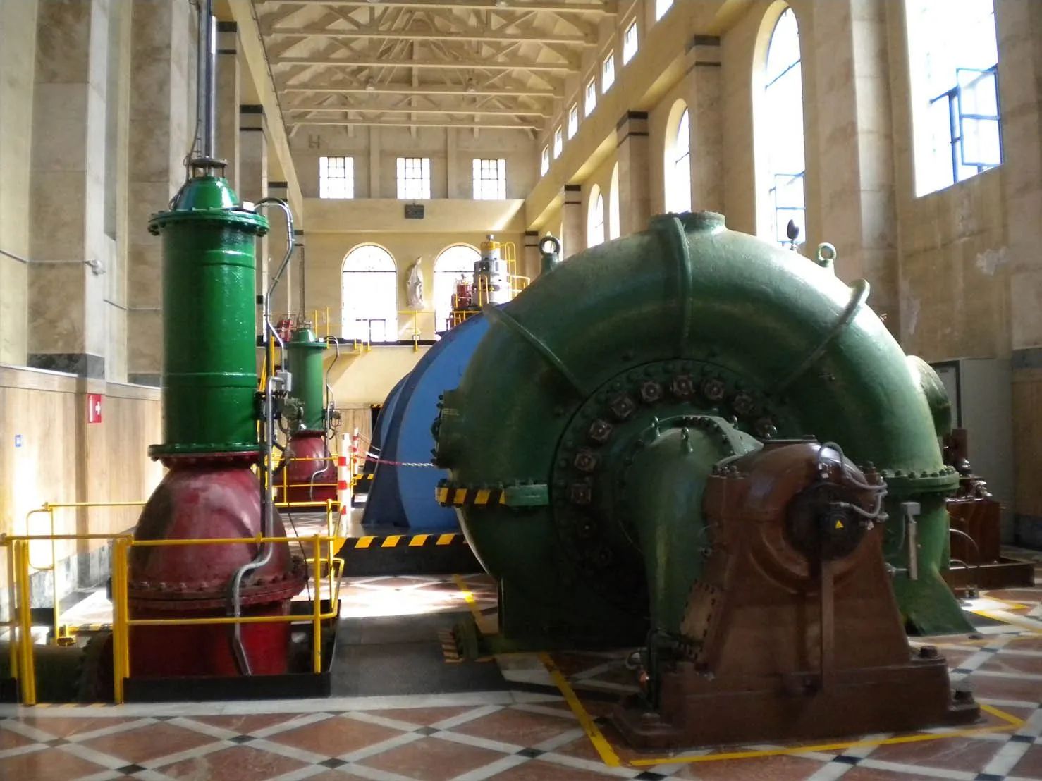 Maquinaria de gran tamaño, verde y roja, en una nave industrial con ventanas arqueadas; probablemente se trate de una central eléctrica.